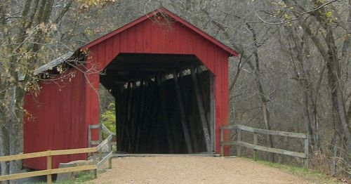 A red covered bridge surrounded by bare trees and a gravel path, with wooden railings on either side.