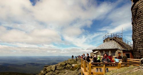 A group of people on a viewing platform at a mountain summit, with a scenic landscape and cloudy sky in the background.