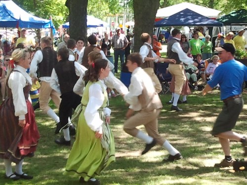 A lively outdoor festival scene with people in traditional costumes dancing, surrounded by spectators and colorful tents.