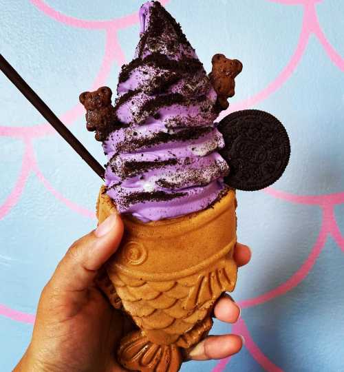 A hand holding a fish-shaped waffle cone filled with purple soft serve, topped with crushed cookies and a chocolate cookie.