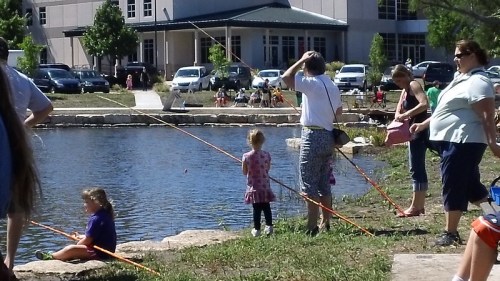 A group of people fishing by a pond, with children and adults enjoying a sunny day outdoors.