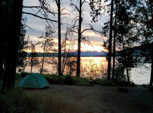 A green tent by a lake at sunset, surrounded by trees and mountains in the background.