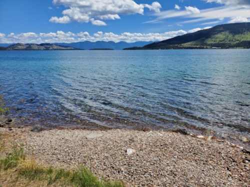 A serene lake view with clear blue water, rocky shore, and mountains under a partly cloudy sky.
