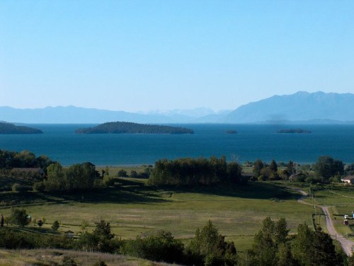 A serene landscape featuring a calm lake with islands, surrounded by green fields and distant mountains under a clear blue sky.