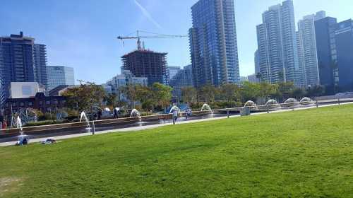 A sunny urban park with green grass, fountains, and tall buildings in the background.