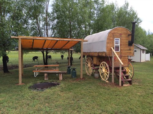 A rustic wooden wagon with a chimney beside a picnic area and fire pit, set in a grassy field with trees and cows in the background.