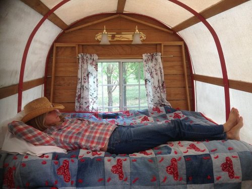 A woman in a cowboy hat relaxes on a bed in a cozy, rustic trailer with floral curtains and a colorful blanket.