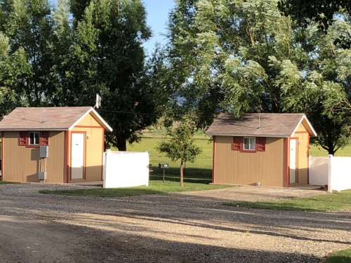 Two small, charming cabins with red shutters, surrounded by green trees and a gravel path.