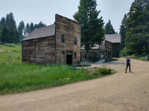 A dusty road leads to old wooden buildings surrounded by trees and grass in a rural setting.