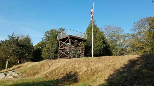 A wooden observation tower with a flagpole stands on a grassy hill, surrounded by trees under a clear blue sky.