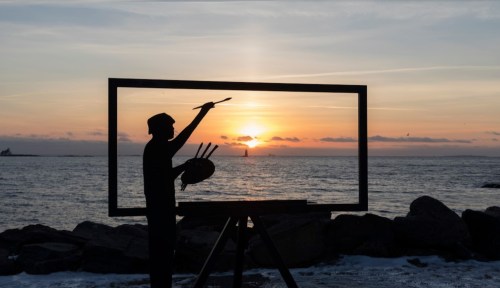 A silhouette of a painter in front of a large frame, capturing a sunset over the ocean.