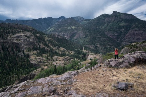 A person in a red jacket stands on a rocky outcrop, overlooking a lush, mountainous landscape under a cloudy sky.