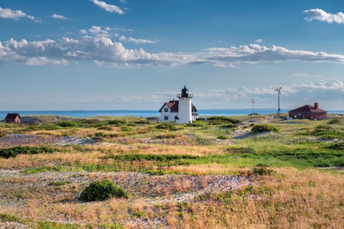 A scenic view of a lighthouse surrounded by grassy dunes and a calm sea under a blue sky with scattered clouds.