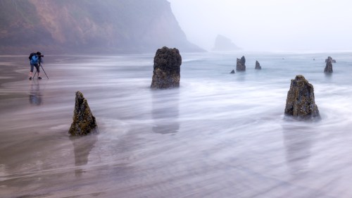 A photographer captures the misty beach scene with rocky formations emerging from the water.