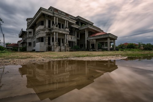 An abandoned, dilapidated building reflected in a puddle, under a cloudy sky.