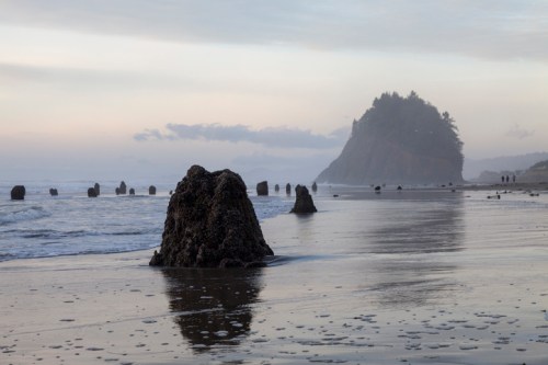 Rocky formations on a sandy beach with gentle waves, under a misty sky and a distant forested hill.
