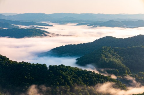 A panoramic view of misty mountains and valleys, with lush greenery and soft clouds blanketing the landscape.
