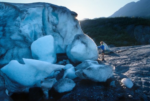 A person sits near large ice formations on a rocky surface, with mountains and a clear sky in the background.