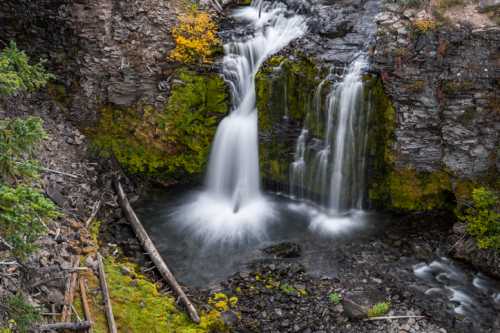 A serene waterfall cascades over rocky terrain, surrounded by lush greenery and autumn foliage.