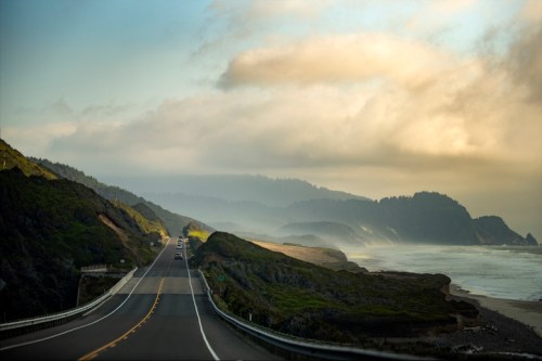 A scenic coastal road winds along cliffs, with misty mountains and ocean waves in the background under a cloudy sky.