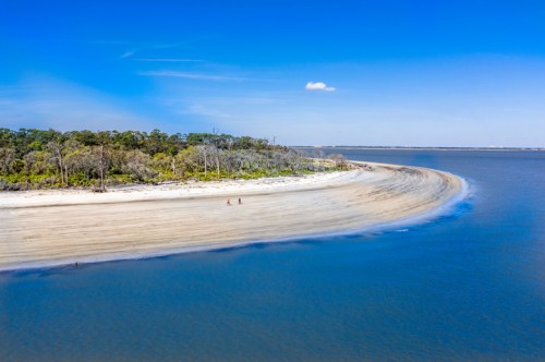 A serene beach with a curved shoreline, surrounded by trees under a clear blue sky. Two people walk along the shore.