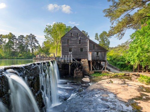 A historic watermill beside a serene river, with a waterfall cascading over rocks and lush greenery surrounding it.