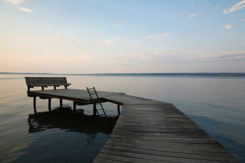 A wooden dock extends over calm water, featuring a bench and a ladder, under a soft, pastel sky.