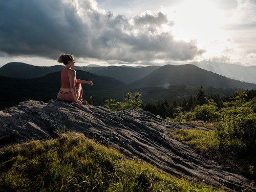 A person sits on a rock, gazing at mountains under a cloudy sky, with sunlight breaking through the clouds.