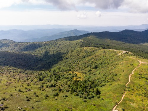 Aerial view of rolling green hills and a winding path, with distant mountains under a partly cloudy sky.