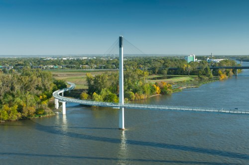 A modern cable-stayed bridge spans a river, surrounded by greenery and urban buildings in the background.
