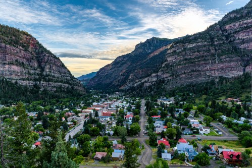 A scenic view of a valley town surrounded by towering mountains and lush greenery under a cloudy sky.