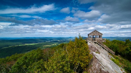 A wooden cabin perched on a rocky outcrop, surrounded by lush green hills and a dramatic sky with scattered clouds.