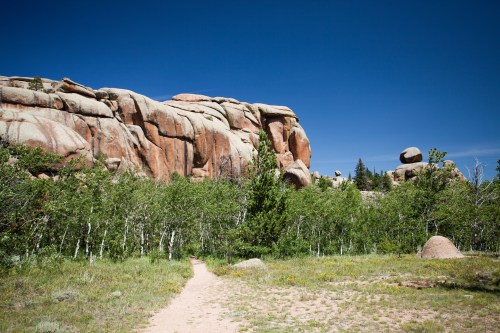 A scenic landscape featuring large rock formations, green trees, and a clear blue sky. A path winds through the foreground.