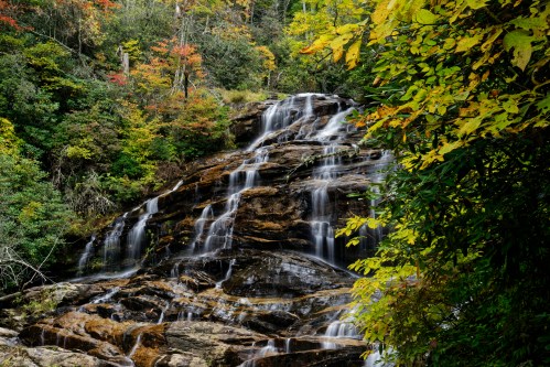 A serene waterfall cascades over rocks, surrounded by vibrant autumn foliage in a lush forest.