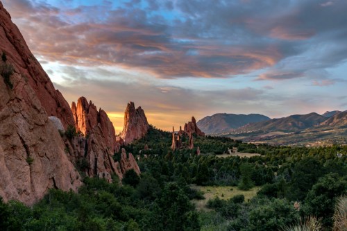 Sunset over rugged rock formations and lush greenery, with mountains in the background and a colorful sky.