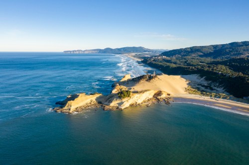 Aerial view of a coastal landscape with sandy cliffs, waves, and a distant shoreline under a clear blue sky.