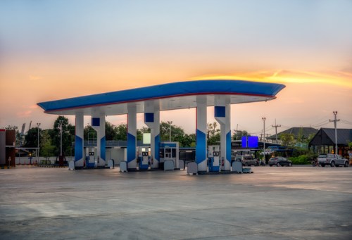A gas station with multiple pumps under a colorful sunset sky, surrounded by trees and parked cars.