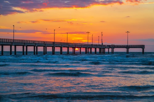 A pier extends over calm waters at sunset, with vibrant orange and purple hues in the sky.