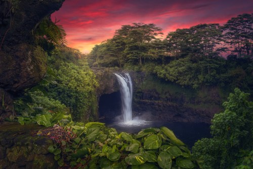 A serene waterfall cascades into a lush green pool, surrounded by vibrant foliage and a colorful sunset sky.