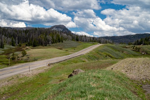 A winding road through lush green hills under a partly cloudy sky, with distant mountains in the background.