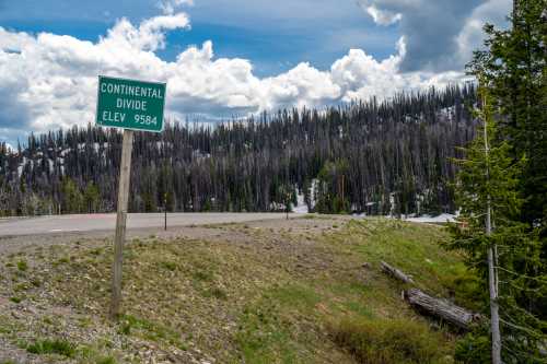 Sign marking the Continental Divide at an elevation of 9,584 feet, with a backdrop of trees and cloudy skies.