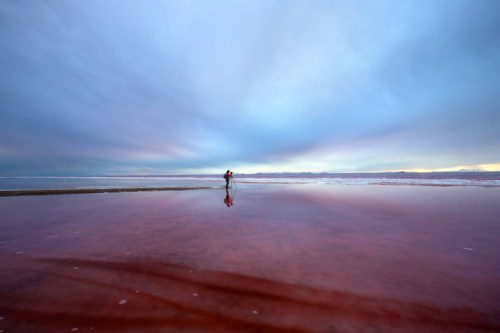 A person stands on a reflective, reddish salt flat under a cloudy sky, creating a serene and expansive landscape.