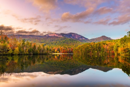 A serene lake reflects colorful autumn trees and mountains under a cloudy sky at sunset.
