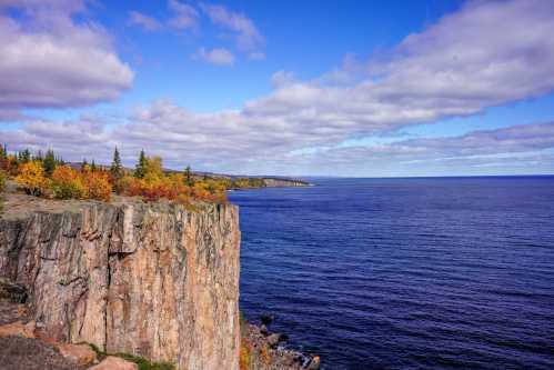 A scenic view of a rocky cliff overlooking a calm blue lake, surrounded by autumn foliage and a cloudy sky.