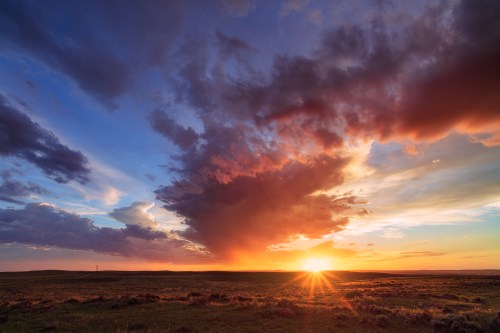 A vibrant sunset over a grassy landscape, with dramatic clouds and warm colors illuminating the sky.