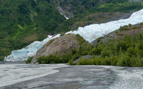 A glacier flows down a rocky landscape, surrounded by greenery and mountains under a cloudy sky.