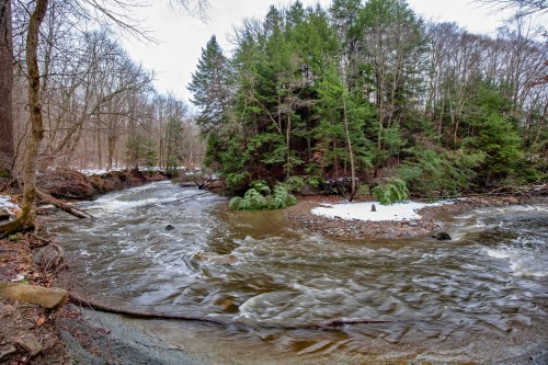 A winding river flows through a forested area, with snow-dusted ground and greenery along the banks.