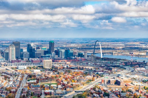 Aerial view of a city skyline featuring tall buildings, a river, and a prominent arch structure under a cloudy sky.