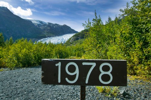 A wooden sign with the year "1978" stands in front of a glacier and lush greenery under a blue sky.