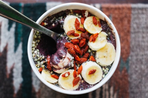 A bowl of smoothie topped with banana slices, goji berries, and hemp seeds, with a spoon resting beside it.